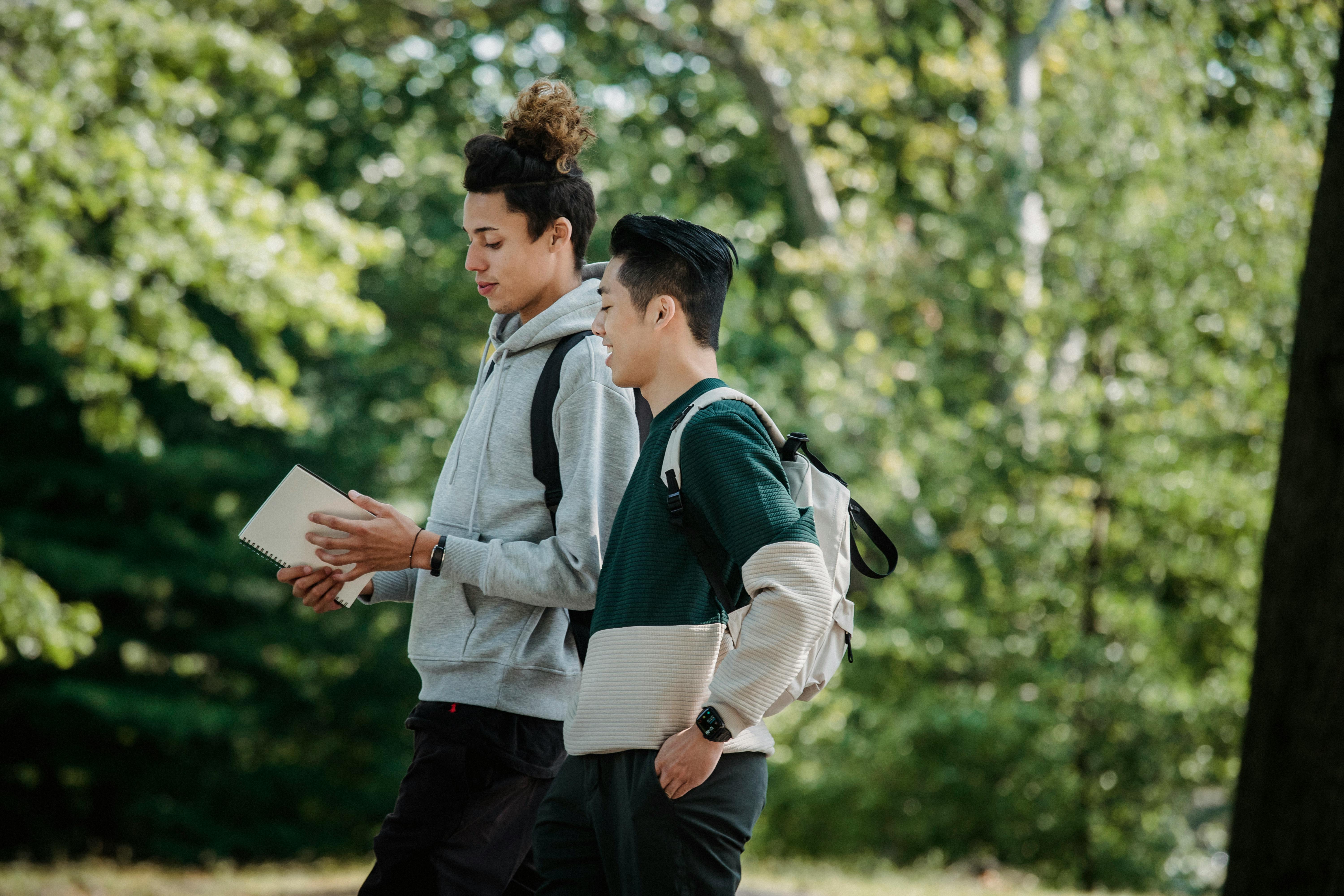 Two young male students with backpacks and notepad walking and discussing in a lush green park.